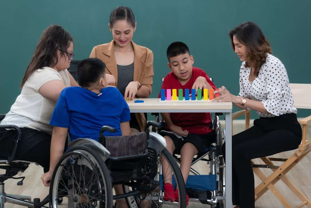 A teacher and an instructional assistant work at a classroom table with a small group of students, including students who use wheelchairs.