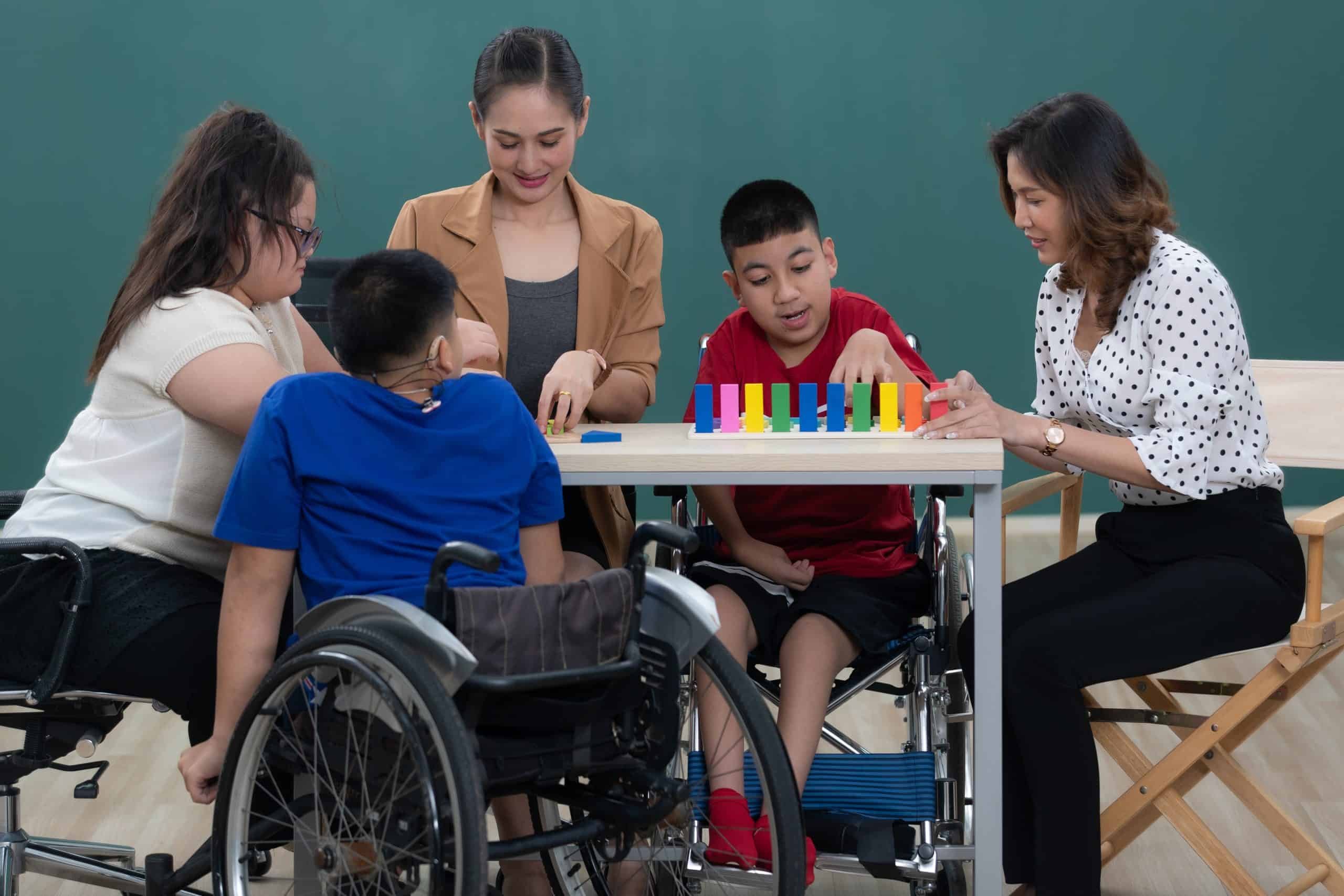 A teacher and an instructional assistant work at a classroom table with a small group of students, including students who use wheelchairs.